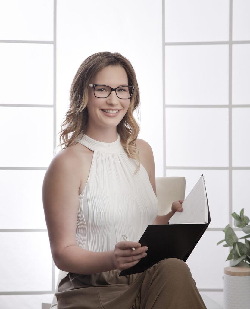 A smiling woman with glasses and a sleeveless white top sits holding an open notebook and pen, capturing a modern business image. She is seated near a potted plant with a bright, softly lit background. - Jodi O Photography