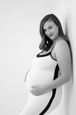 A pregnant woman in a sleeveless, form-fitting maternity dress with dark side panels stands sideways, gently cradling her belly and smiling at the camera against a plain white background. - Jodi O Photography