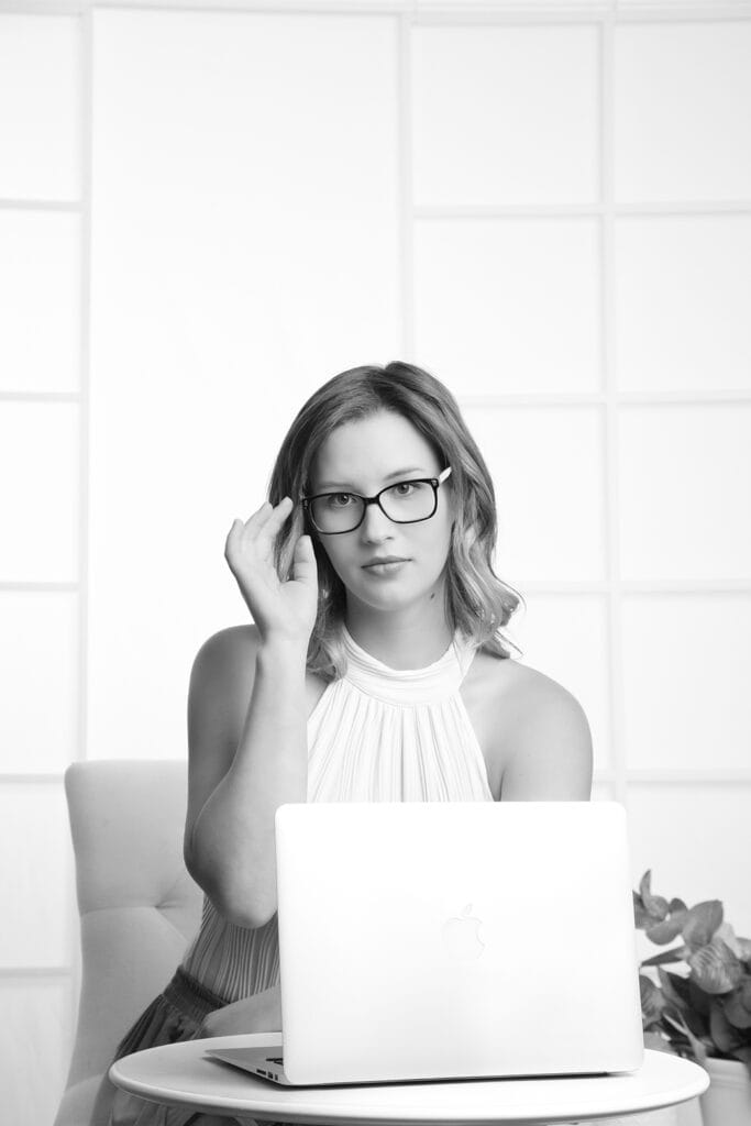 A woman wearing glasses and a sleeveless top sits at a table with an open laptop, embodying a modern business image. She looks at the camera, adjusting her glasses, set against a bright and minimalistic background. - Jodi O Photography