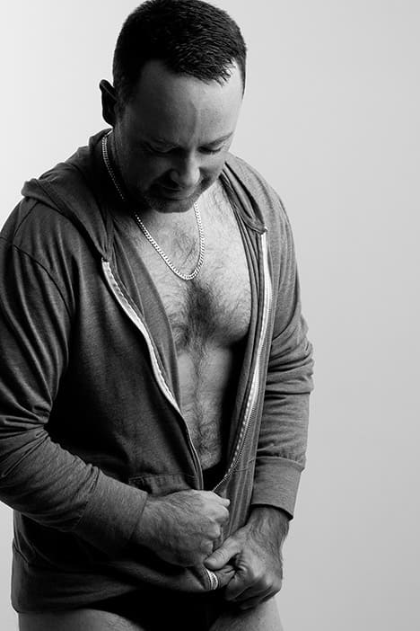 A man in a partially unzipped hoodie exposing his chest looks down while pulling at the zipper. This black and white Men’s Boudoir photo, set against a neutral background, captures a bold moment of vulnerability—perfect inspiration for Calgary boudoir photography. - Jodi O Photography
