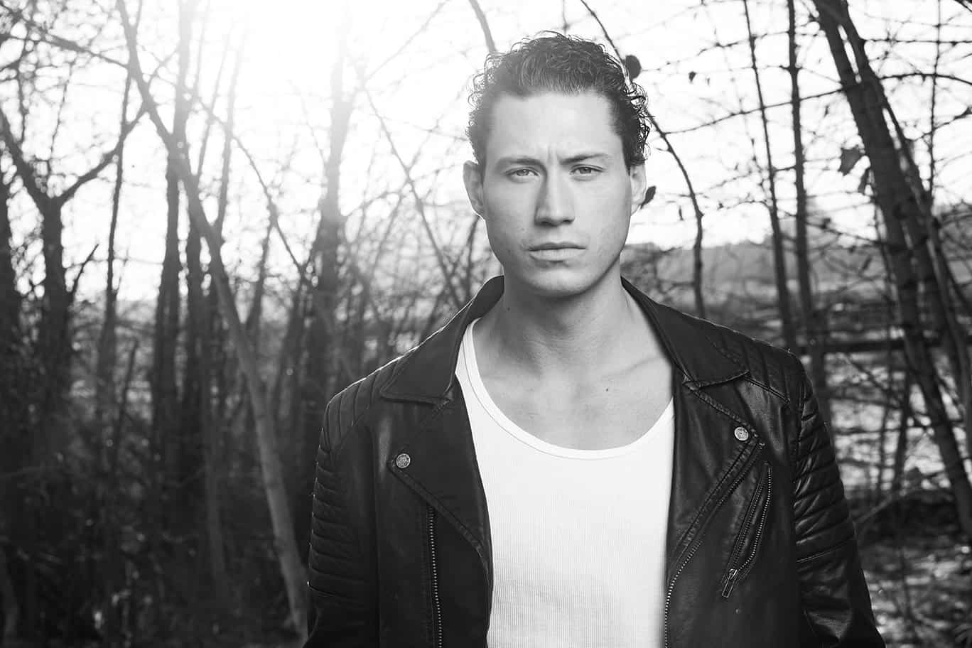 A young man with short curly hair wearing a black leather jacket and white shirt stands outdoors in front of leafless trees, sunlight shining through the branches. This black and white shot is perfect for actor headshots Calgary style. - Jodi O Photography