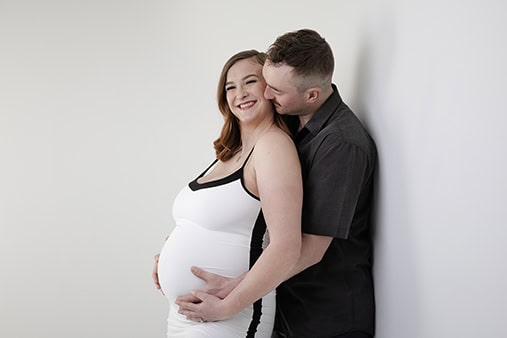 A smiling pregnant woman in a white dress stands against a white wall during a joyful maternity moment as a man hugs her from behind, resting his hands on her belly and kissing her cheek. - Jodi O Photography