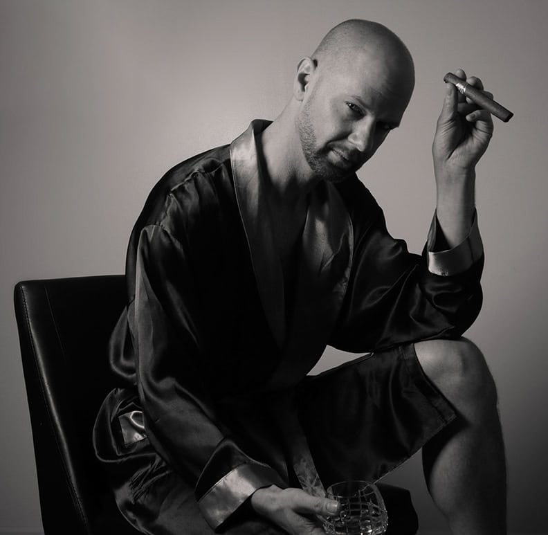 A bald man in a satin robe sits on a chair, holding a cigar and a glass, gazing at the camera with a relaxed expression. This black and white photo captures men’s boudoir photography style, perfect for modern Calgary gents. - Jodi O Photography