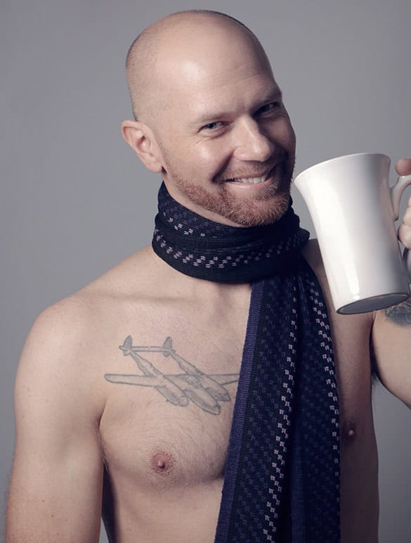 A smiling bald man with a reddish beard wears a dark patterned scarf and holds a white mug. Shot in Calgary, this men’s boudoir portrait features him shirtless, revealing an airplane tattoo on his chest against a plain background. - Jodi O Photography