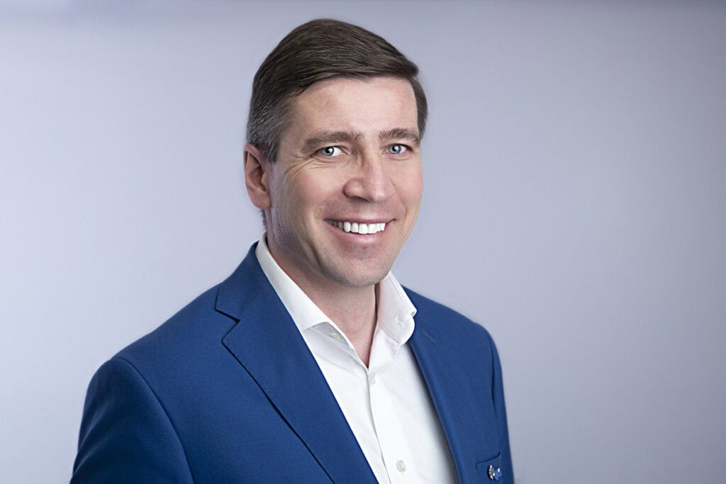 A man wearing a blue suit jacket and white shirt, with a pop of style and modern confidence, smiles at the camera against a plain light gray background. - Jodi O Photography