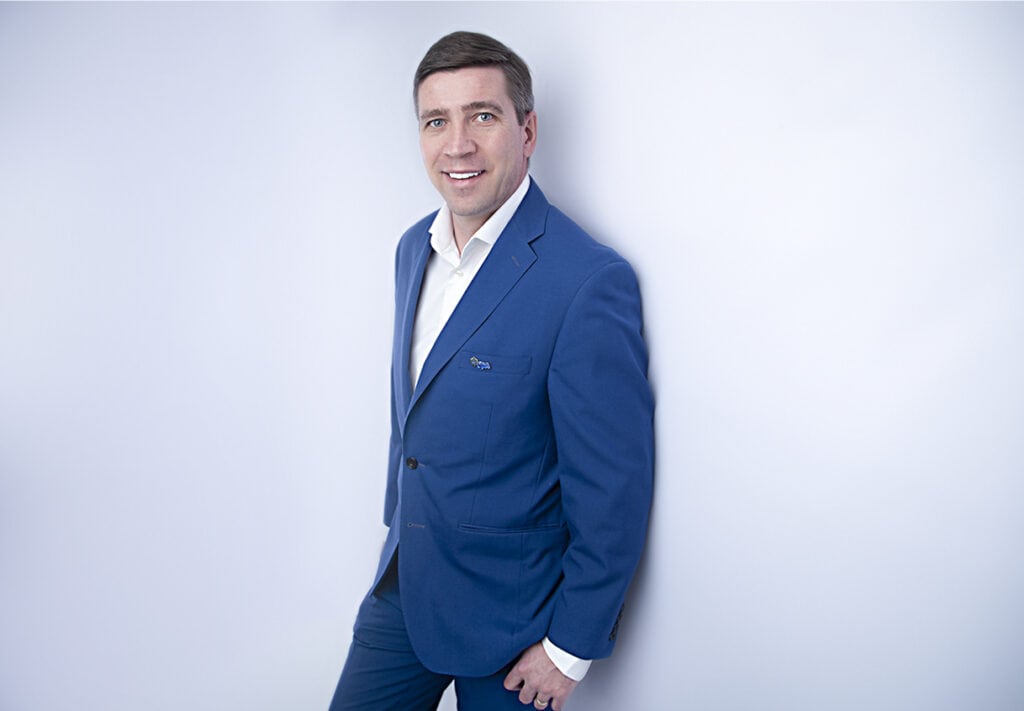 A man in a blue suit and white shirt stands against a plain light background, smiling at the camera with one hand in his pocket, radiating modern confidence and a pop of style. - Jodi O Photography