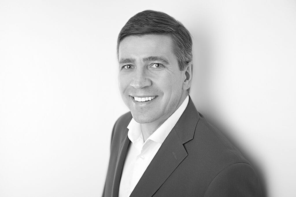 Black-and-white portrait of a smiling man in a suit jacket and white shirt, standing against a plain light background, looking slightly to the side with modern confidence and a subtle pop of style. - Jodi O Photography