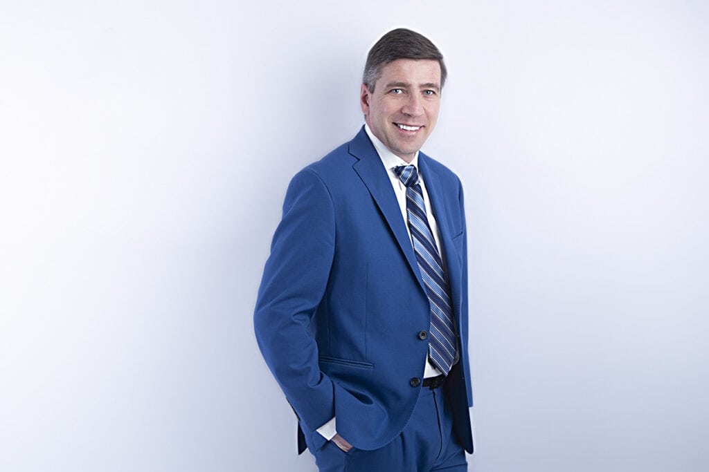 A man wearing a blue suit with a striped tie stands against a plain light background, smiling with one hand in his pocket—radiating modern confidence and a pop of style. - Jodi O Photography