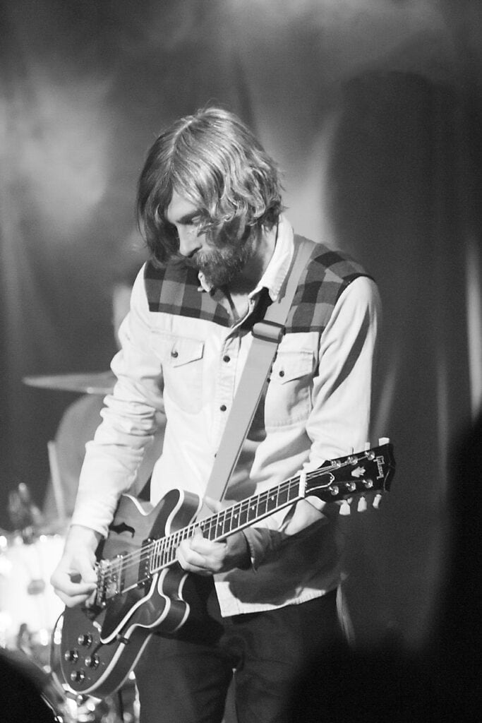 A man with medium-length hair and a beard plays an electric guitar on stage. He is wearing a light-colored shirt with a checkered pattern on the shoulders. The dimly lit background and visible drum set capture dynamic Music Photography. - Jodi O Photography