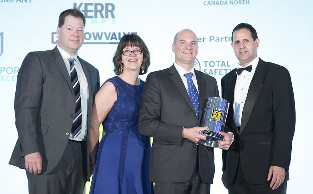 Four people in formal attire stand together at an award event, with one man holding a trophy. They are smiling, and sponsor logos are visible on the backdrop—a classic moment captured by a Calgary Corporate Event Photographer. - Jodi O Photography