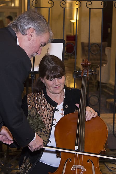 A man shows a woman how to play a cello, holding the bow together. The woman sits holding the cello, while the man leans in to guide her hand. This intimate moment of music photography is captured indoors in a warmly lit room. - Jodi O Photography