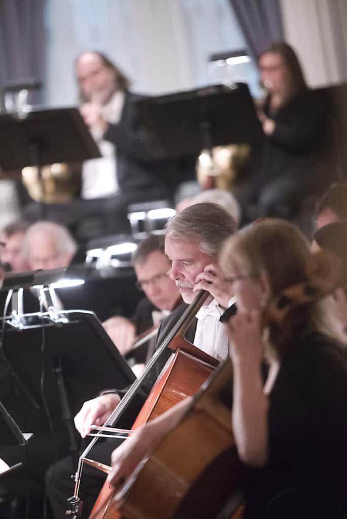 Musicians play string instruments in an orchestra. A man focused on his cello is in the foreground, while other musicians perform with sheet music in the background—capturing a moment of music photography under soft indoor lighting. - Jodi O Photography