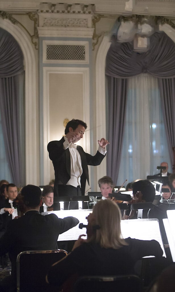 A conductor in formal attire leads an orchestra during a performance in an elegant, classical concert hall with tall windows and elaborate curtains. Music photography captures musicians playing string instruments in the foreground. - Jodi O Photography