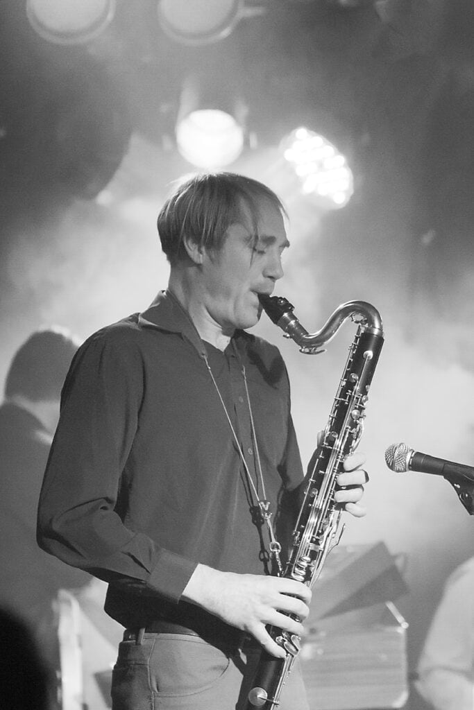 A musician playing a bass clarinet on stage, illuminated by stage lights, with a microphone nearby. The smoky, blurred background captures the vibrant energy of live music photography. - Jodi O Photography