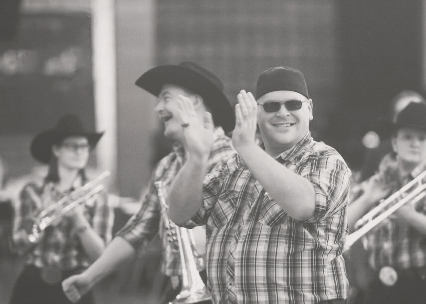 A group of people in plaid shirts and cowboy hats play saxophones and clap while participating in a parade or outdoor event. Perfect for Music Photography, the main person in front is smiling, wearing sunglasses. The black-and-white image captures lively moments. - Jodi O Photography