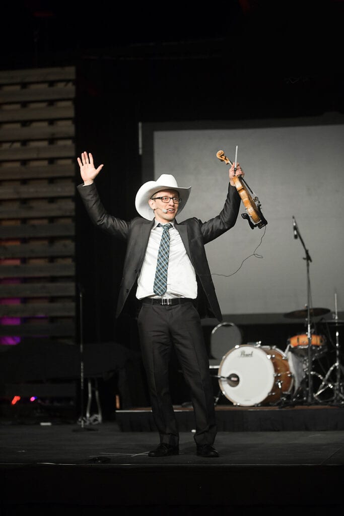 A man in a suit and cowboy hat stands on stage with arms raised, holding a violin in one hand. Capturing the excitement of live music photography, drums and a large display screen are visible in the background. - Jodi O Photography