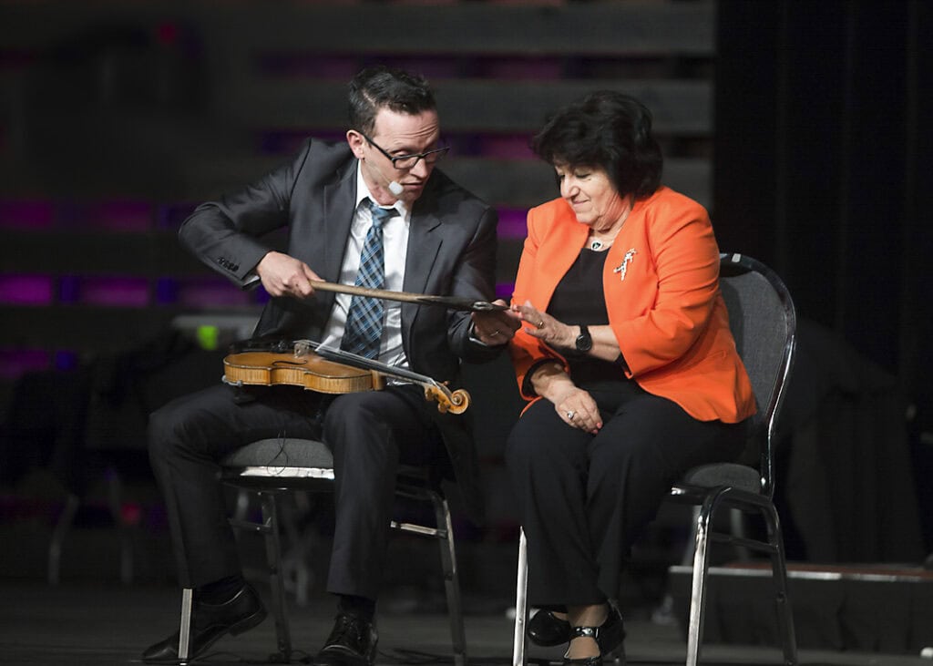 A man in a suit shows a violin bow to a seated woman in an orange blazer during an event on stage. Capturing this candid moment of conversation, the image reflects the essence of Event Photography. - Jodi O Photography