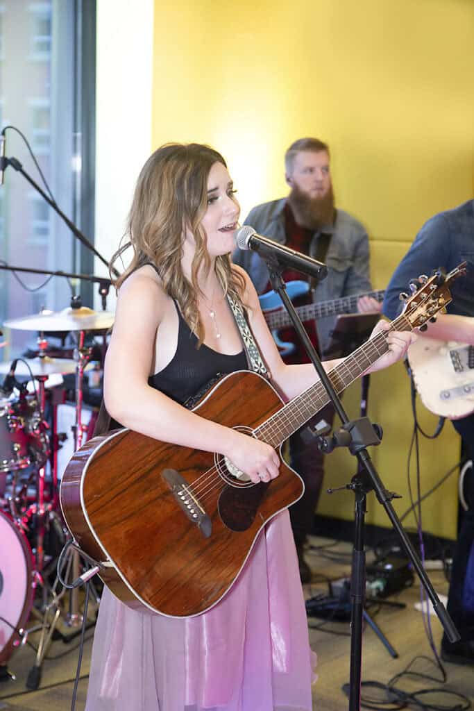 A young woman in a black top and pink skirt sings and plays acoustic guitar into a microphone. Captured through music photography, behind her, a man plays bass and another person is on electric guitar, with a drum set in the background. - Jodi O Photography