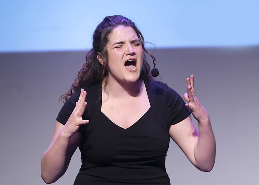 A woman with curly hair wearing a black dress and headset microphone passionately sings or speaks on stage, her eyes closed and hands raised, captured in striking detail—perfect for event photography or music photography enthusiasts. - Jodi O Photography