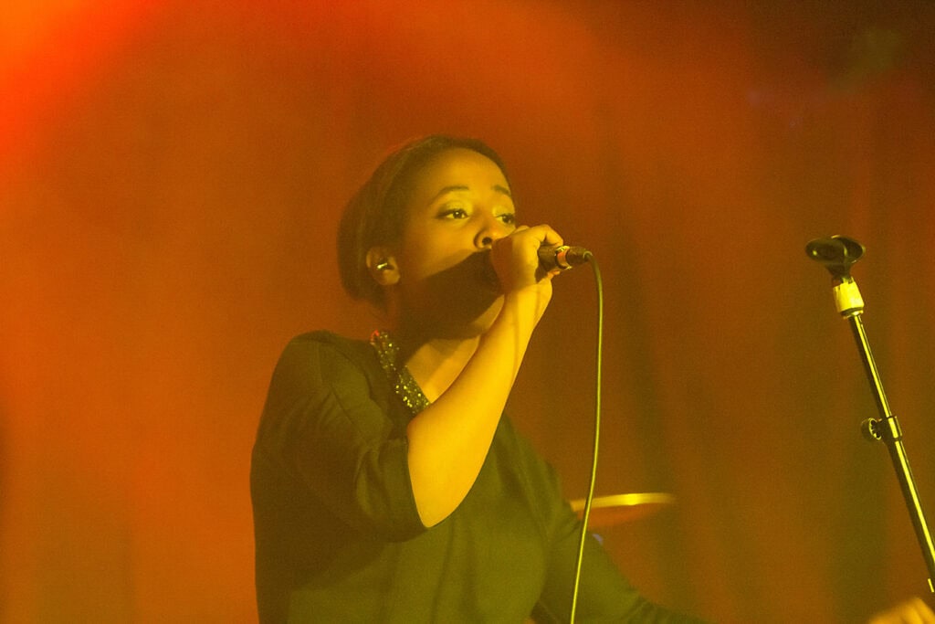 A singer in a black outfit performs on stage, holding a microphone close to her mouth as warm yellow and orange stage lighting creates a dramatic effect—an impressive example of event photography. - Jodi O Photography