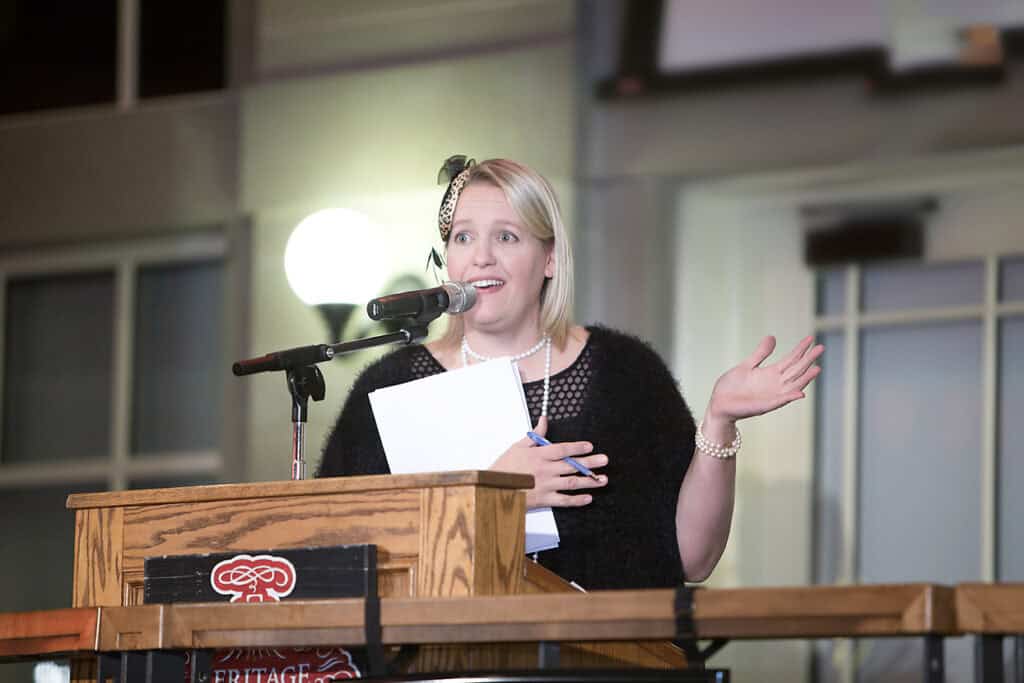 A woman with blonde hair speaks enthusiastically at a podium, holding papers in one hand and gesturing with the other at a business event. She wears a black outfit with pearls in a well-lit indoor setting—captured by a Calgary corporate event photographer. - Jodi O Photography