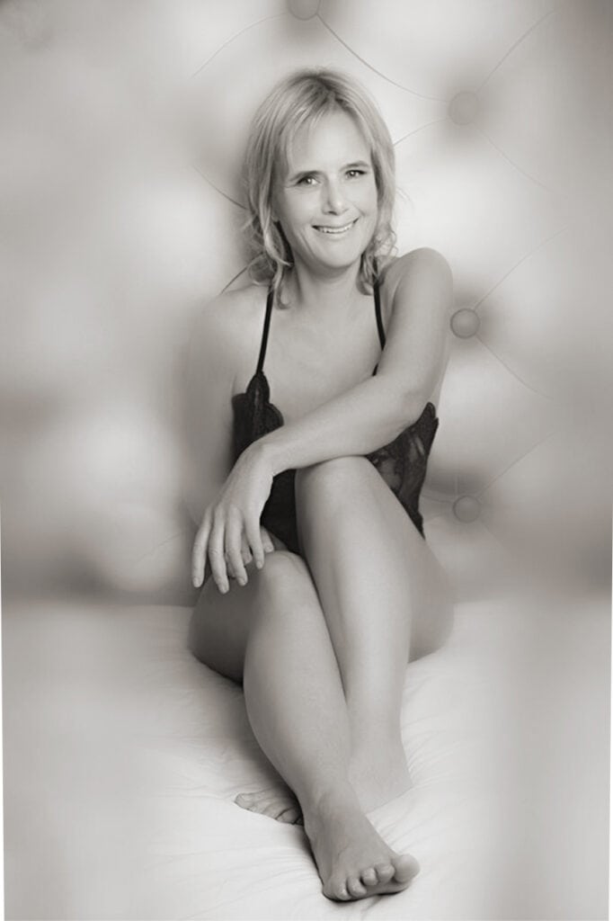 A woman with blonde hair sits on a bed, smiling and wearing a dark, strappy top. The photo is in black and white, with a soft, blurred background. She sits with her legs crossed and one arm resting on her knee. - Jodi O Photography