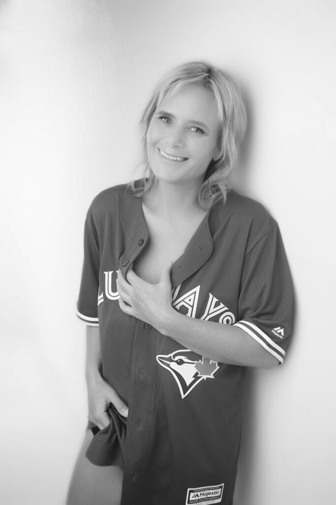 A smiling woman with light hair wears an oversized Toronto Blue Jays jersey, holding it closed at the front. The image is in black and white with a soft, blurred background. - Jodi O Photography