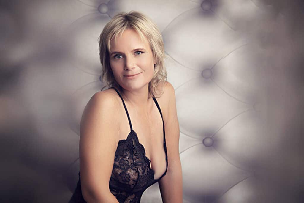 A woman with short blonde hair wearing a black lace dress sits against a soft, tufted background, smiling gently at the camera. - Jodi O Photography