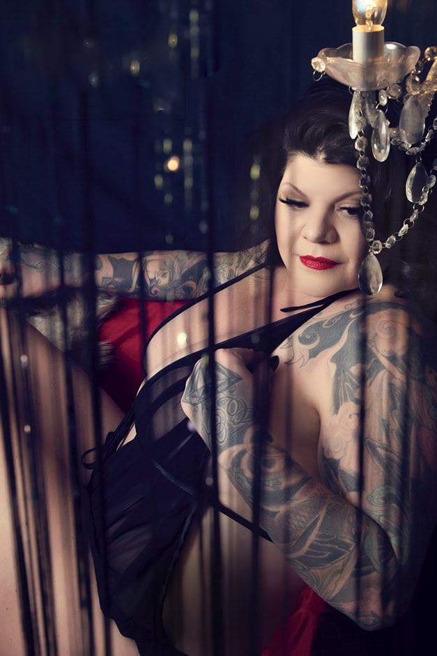 A woman with long dark hair, bright red lipstick, and extensive tattoos poses in sheer black lingerie under a chandelier, with dramatic lighting and black cords in the foreground. - Jodi O Photography