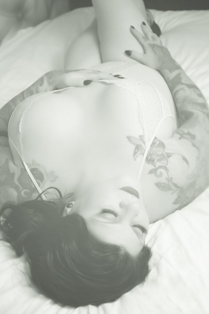 A woman with tattoos lies on her back on a bed, wearing light-colored lingerie. Her hand rests on her torso, and her eyes are closed. The photo has a soft, muted, and dreamy tone. - Jodi O Photography