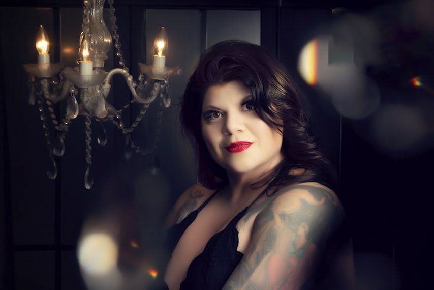 A woman with long dark hair and red lipstick, showing colorful tattoos on her arm, poses in front of a black background with a lit crystal chandelier to her left. The lighting is soft and glamorous. - Jodi O Photography