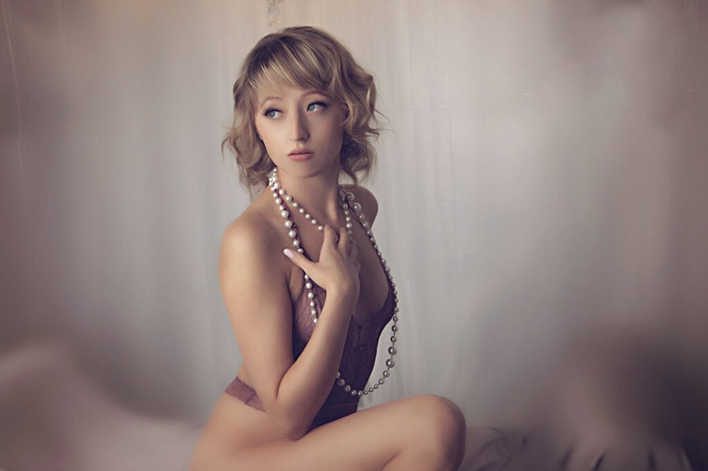 A woman with short, wavy blonde hair poses in a mauve lingerie bodysuit, sitting sideways and looking off-camera. She holds a long pearl necklace against her chest, with a soft, blurred background. - Jodi O Photography