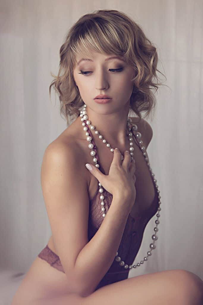 A woman with short, wavy blonde hair wearing a mauve lace top and a long pearl necklace gazes downward with a soft expression, her hand resting on her chest. The background is softly blurred. - Jodi O Photography