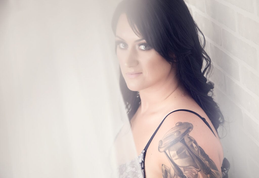 A woman with long dark hair and strong ink on her upper arm gazes softly at the camera, standing beside a white brick wall with a sheer curtain and hints of lace partially obscuring the view. - Jodi O Photography