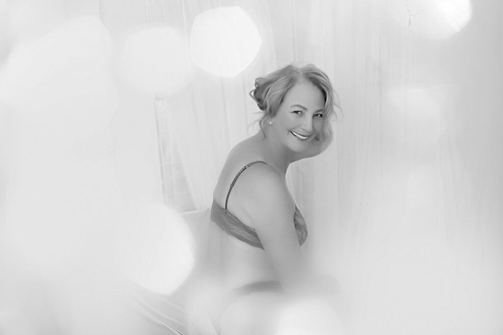 A woman with short light hair, wearing lingerie, sits on a bed and smiles at the camera. The black and white photo, captured by a Calgary Boudoir Photographer, features soft, blurred light effects for a romantic boudoir feel. - Jodi O Photography