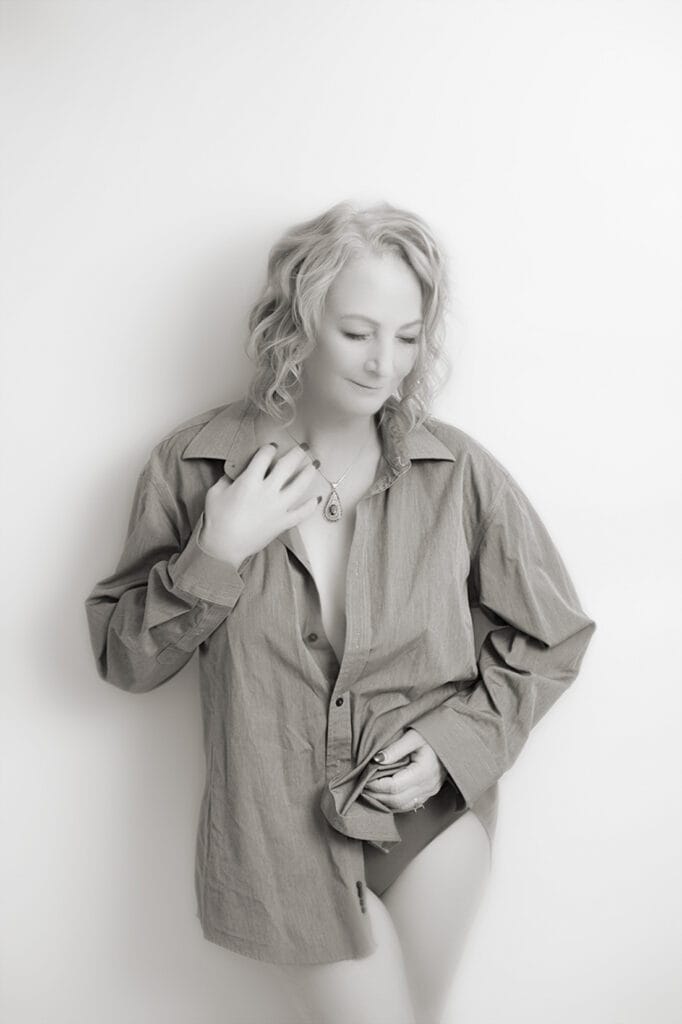 A person with light curly hair stands in a studio against a plain white background, wearing an oversized button-down shirt. They look down with a gentle smile, holding the shirt closed at the waist and touching their necklace. The image is in black and white. - Jodi O Photography