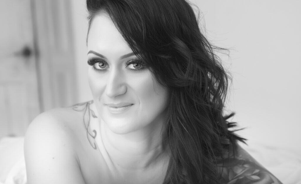A woman with long dark hair and strong ink on her shoulder smiles softly at the camera in a black and white portrait, set against a blurred indoor background—a striking moment from a boudoir session. - Jodi O Photography