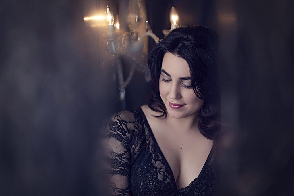 A woman with dark hair in a black lace dress smiles softly while looking down, with a chandelier glowing softly in the background. The scene has a dim, elegant, and intimate atmosphere. - Jodi O Photography