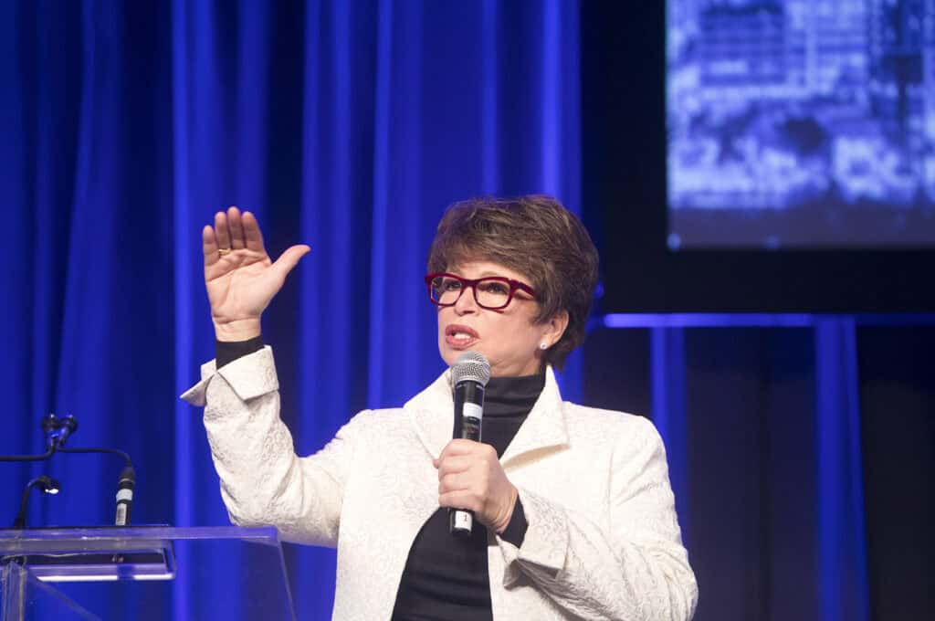A woman in red glasses and a white jacket speaks into a microphone at a podium, gesturing with her left hand. Blue curtains and a screen are visible behind her, capturing dynamic Speakers Photography at Event Moments. - Jodi O Photography