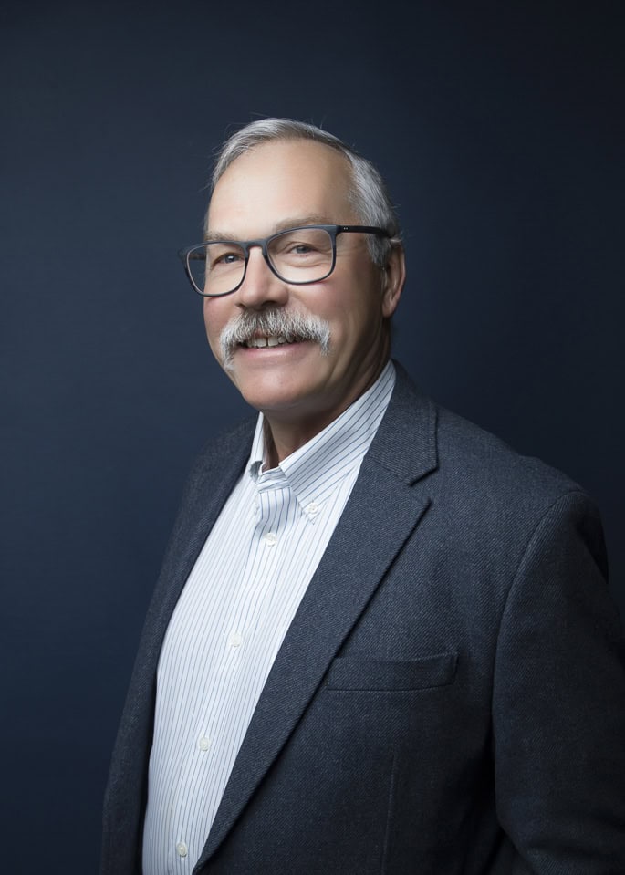 A smiling older man with gray hair, a mustache, and glasses wears a dark blazer over a light striped shirt, bringing sharp style to his look as he poses against a dark blue background. - Jodi O Photography