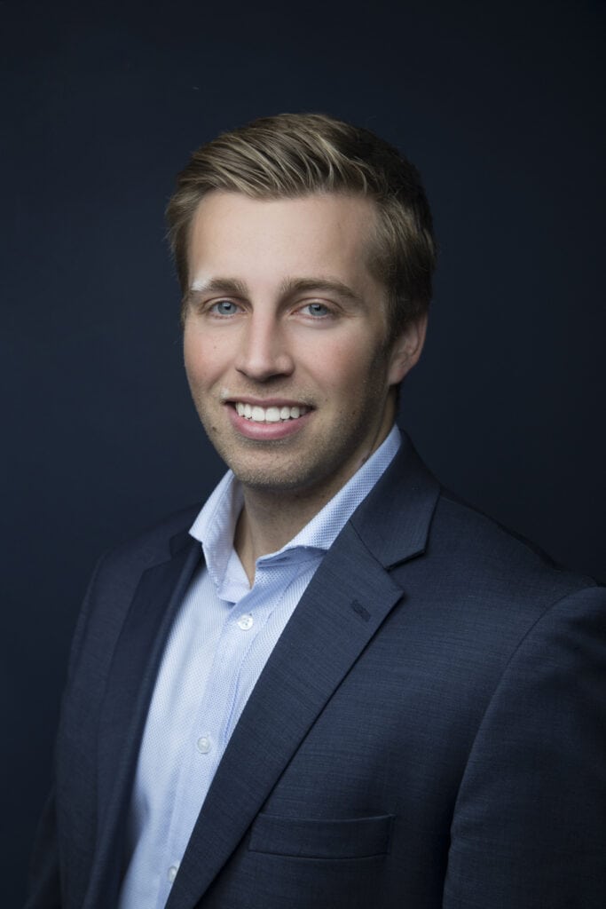 A young man with short, light brown hair wearing a dark suit jacket and light blue shirt smiles against a dark, plain background, capturing a sharp style with a subtle downtown edge. - Jodi O Photography