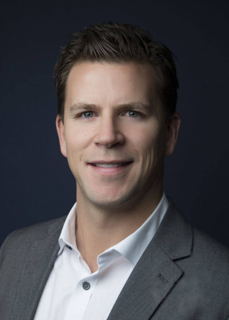 A man with short brown hair wearing a grey suit jacket and white shirt showcases sharp style, smiling slightly in front of a dark background. - Jodi O Photography