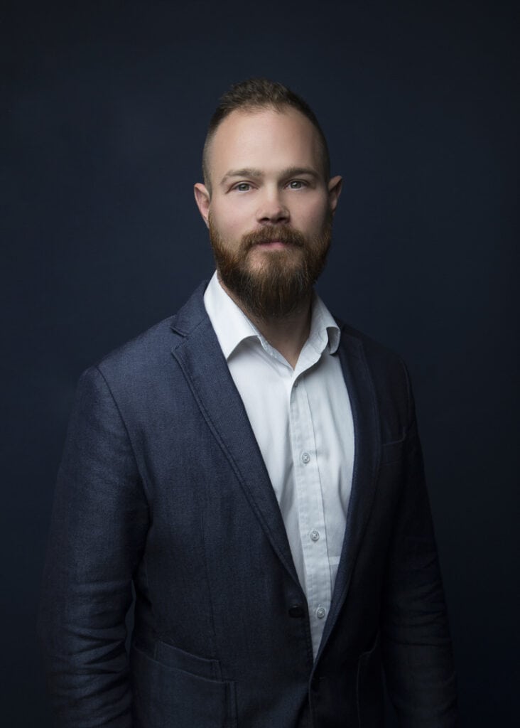 A man with a beard and short hair, dressed in a dark blue blazer and white shirt, stands against a dark background, exuding Sharp Style and Downtown Edge as he looks confidently at the camera. - Jodi O Photography