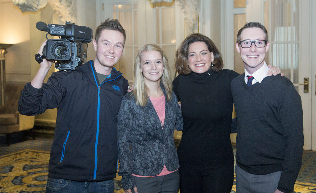 Four people stand together inside a well-lit, ornate room at a business event. One person on the left holds a video camera, and all are smiling at the camera. The group includes two women and two men in business casual attire. - Jodi O Photography