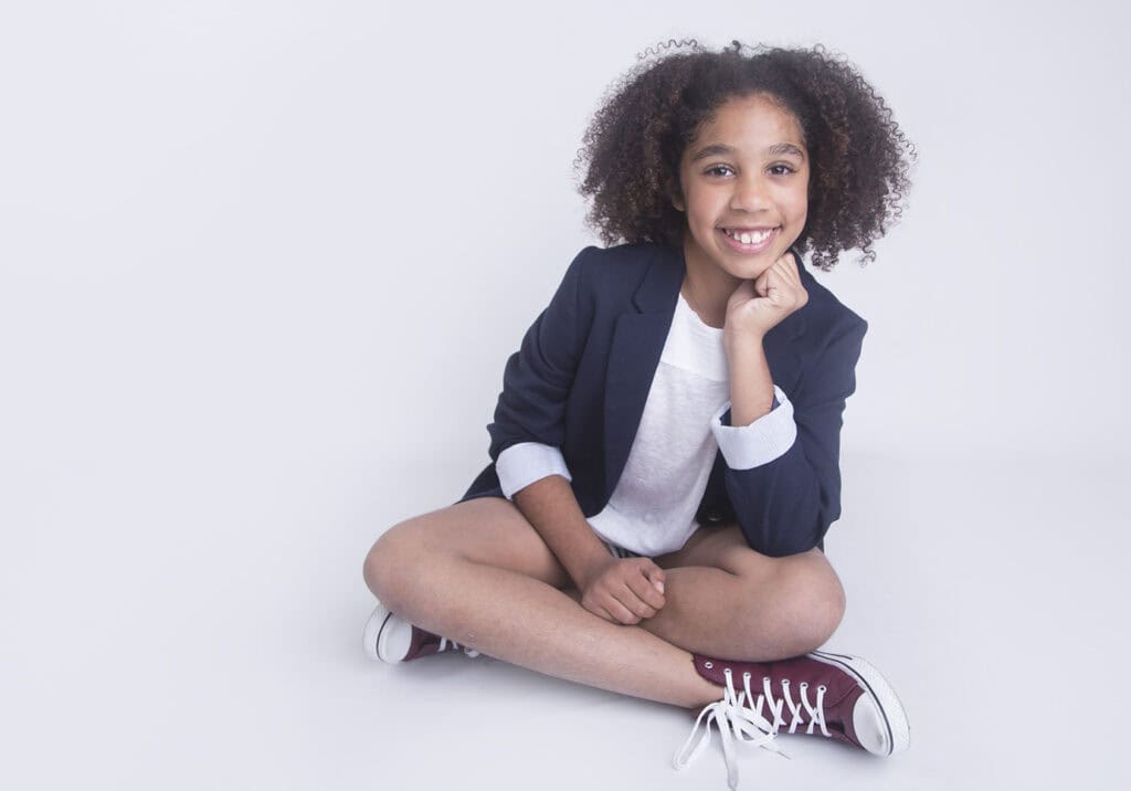 A smiling young girl with curly hair sits cross-legged on the floor, styled for a photography session by a Calgary photographer, wearing a navy blazer, white t-shirt, and maroon sneakers against a plain white background. - Jodi O Photography