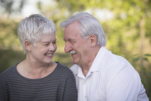 An older couple with gray hair smiles and leans close to each other outdoors, enjoying a happy moment together. The blurred greenery highlights this joyful Calgary photography session. - Jodi O Photography