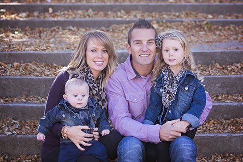 A smiling family of four, including a man, woman, young girl, and baby boy, sit together on outdoor stone steps covered with autumn leaves. Captured by a Family Photographer in Calgary, they wear casual fall clothing and look at the camera. - Jodi O Photography