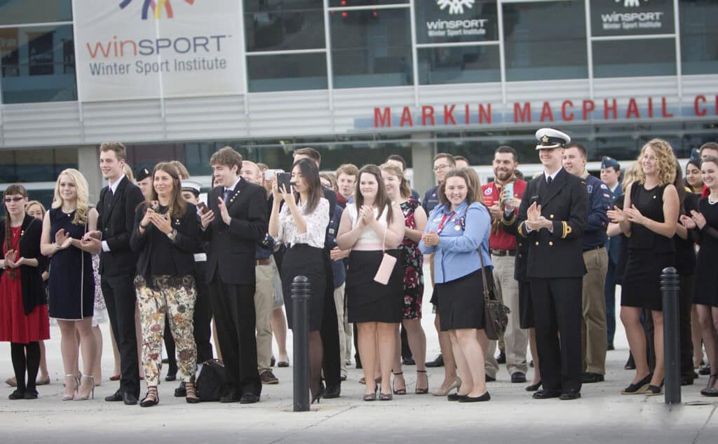 A diverse group of people, dressed in semi-formal and formal attire, stand outside the Markin MacPhail Centre at WinSport, clapping and smiling during a business event captured by a Calgary corporate event photographer. - Jodi O Photography