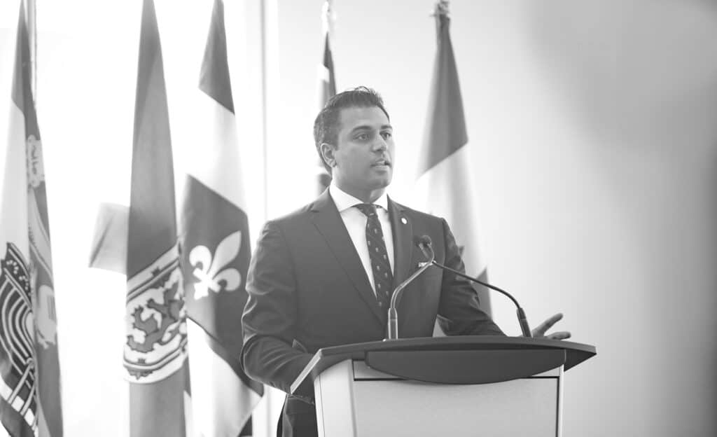 A man in a suit speaks at a podium with a microphone, gesturing with one hand. Several national and provincial flags are displayed in the background, capturing the moment of a Business Event by a Calgary Corporate Event Photographer in black and white. - Jodi O Photography