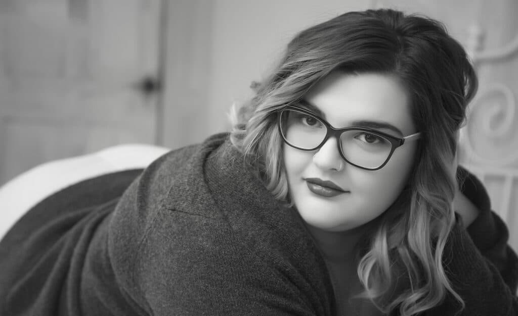 A woman with wavy hair and glasses, wearing a dark sweater, is lying down and looking at the camera with a slight smile. The image is in black and white. - Jodi O Photography